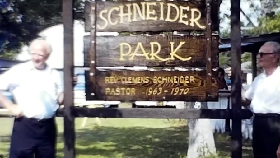 Father Clemens Schneider standing at left beside the carved wooden Schneider Park sign at the 1970 dedication, with a second man at right